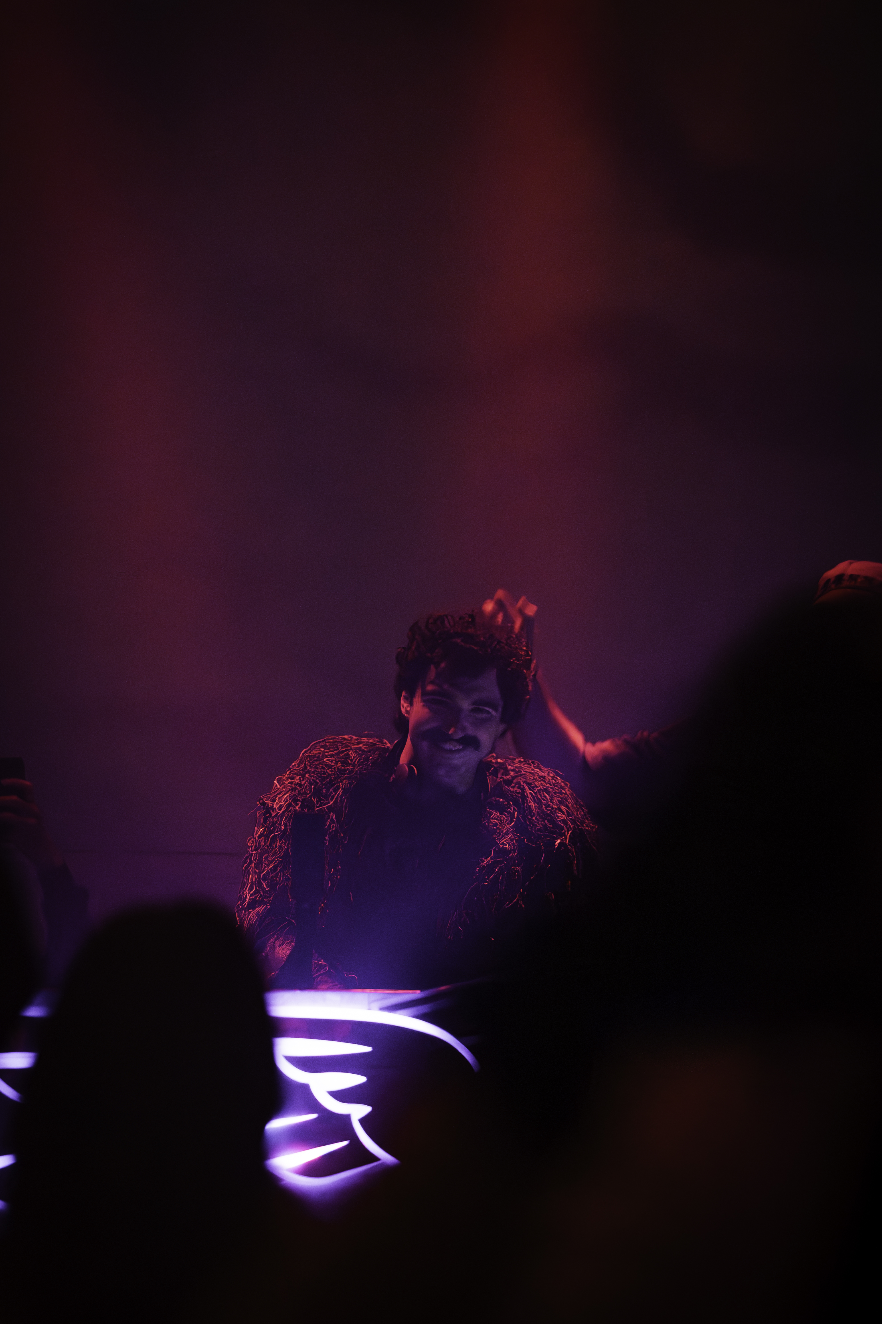 DJ in fringe costume grinning at the decks, purple haze and crowd silhouettes